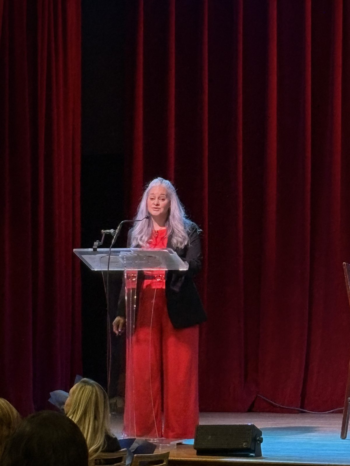 Dalia speaking at a clear podium in front of red stage curtains