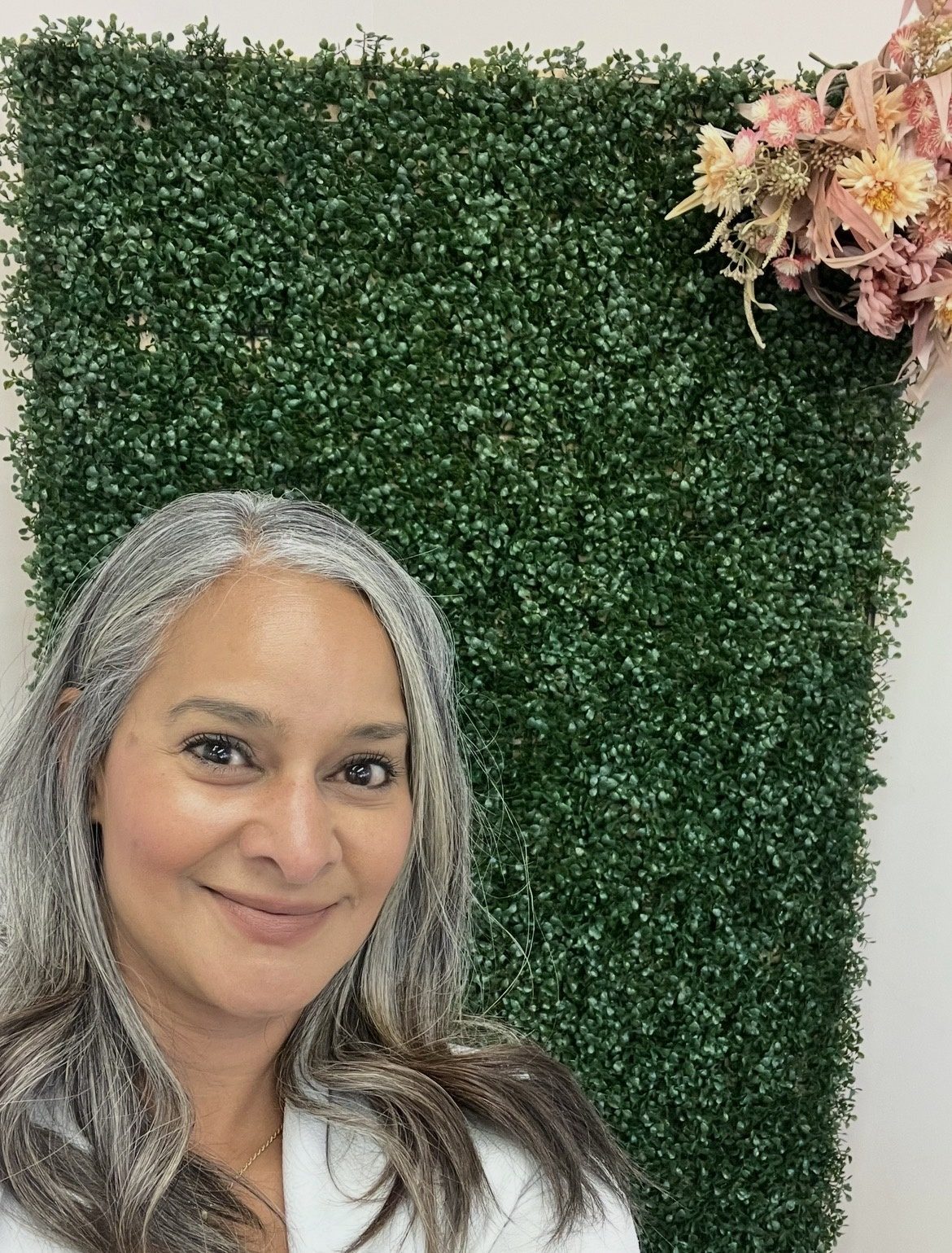 Dalia smiling in front of a green hedge wall with dried flowers